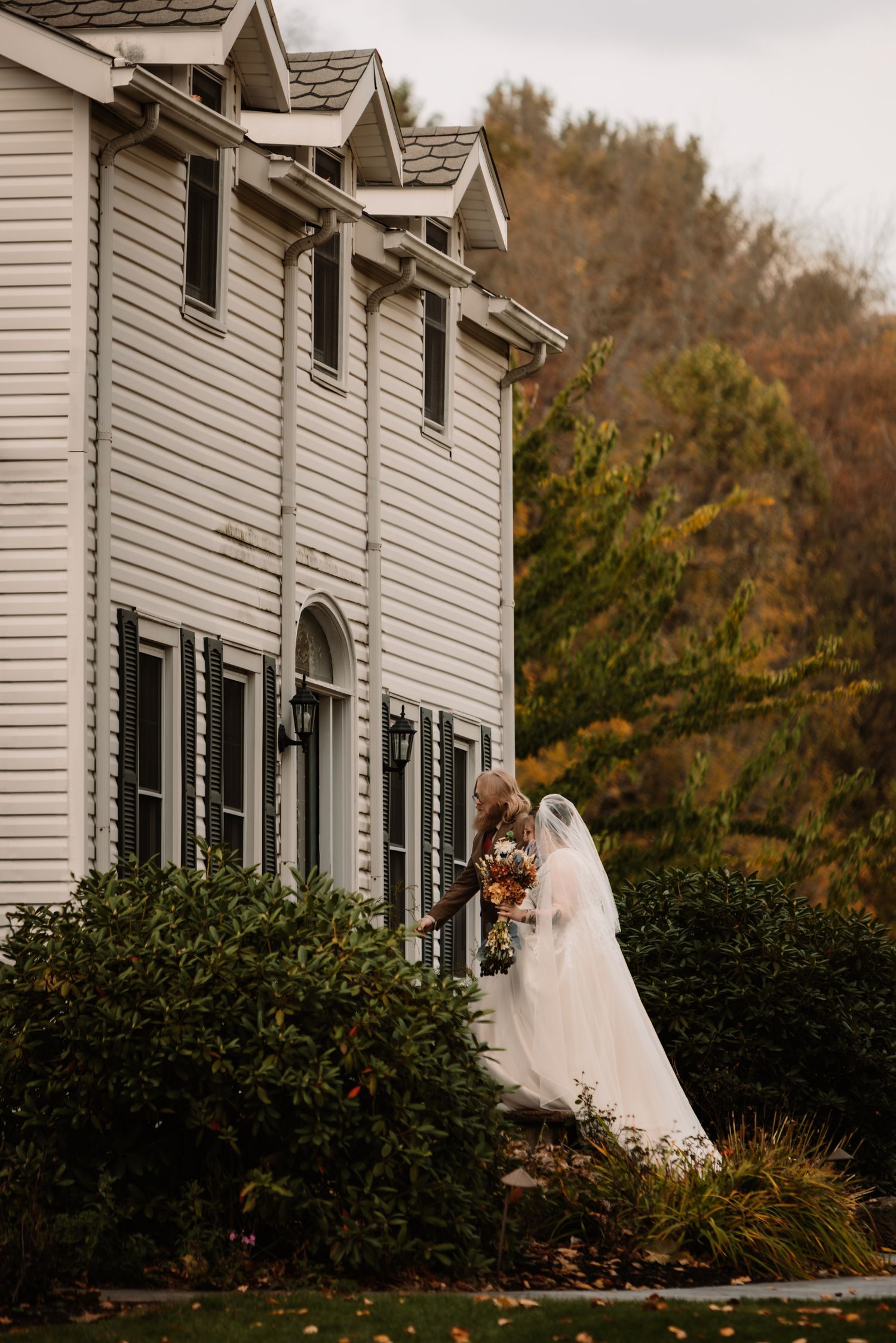 White colonial style house with black shutters serves as backdrop for autumn wedding photography session.