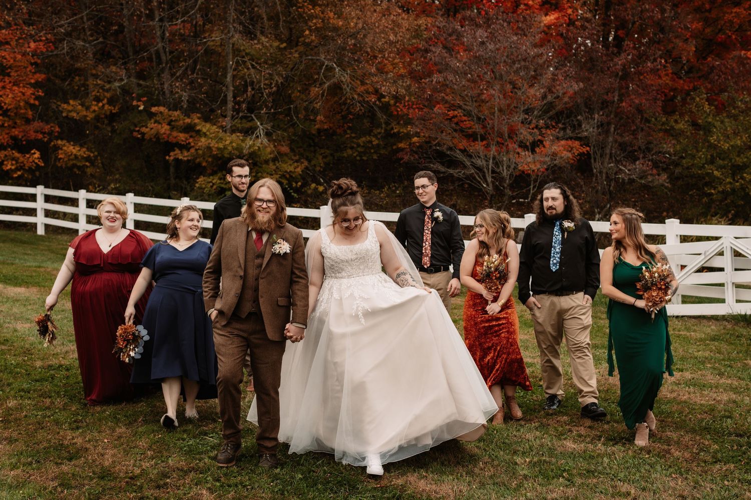 A wedding party walks together on a lawn with autumn foliage and white fences in the background.
