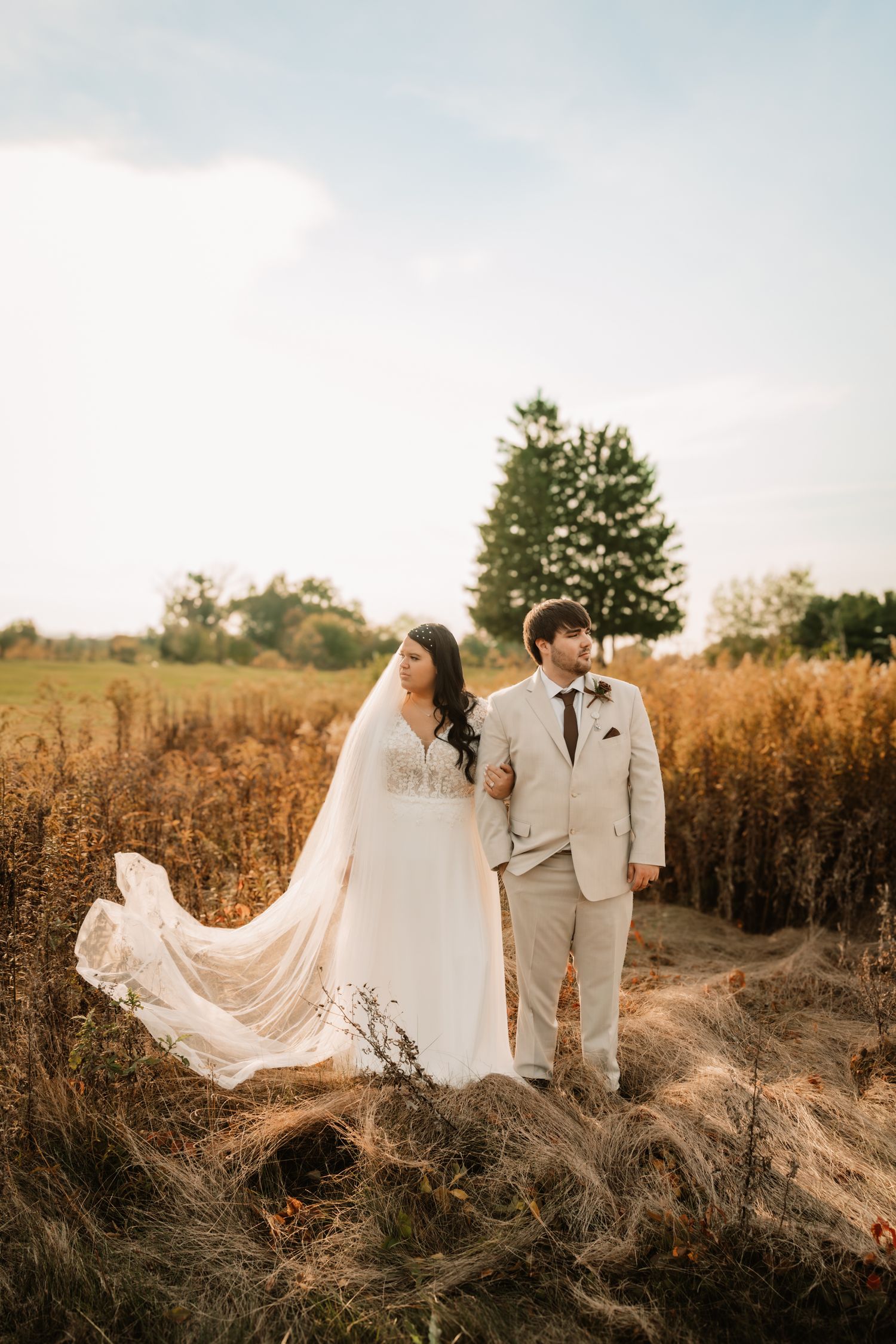A couple in wedding attire embrace in a golden field at sunset as the bride's veil flows dramatically in the wind.