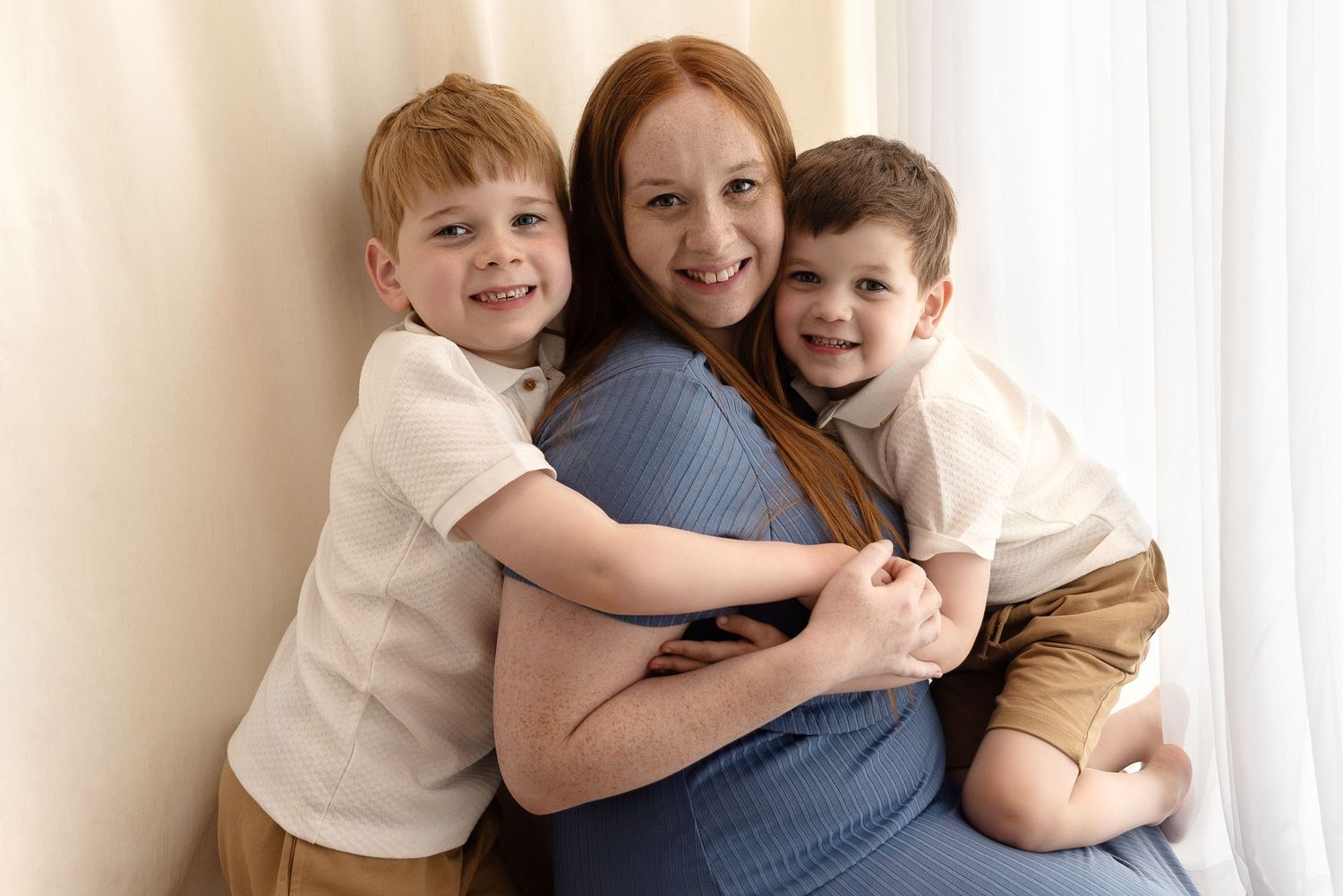 A happy expectant mother wearing a blue dress embraces her two small children against a light background.