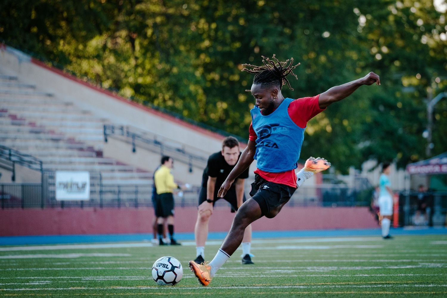 Soccer player with dreadlocks kicking ball on stadium field during evening practice session.