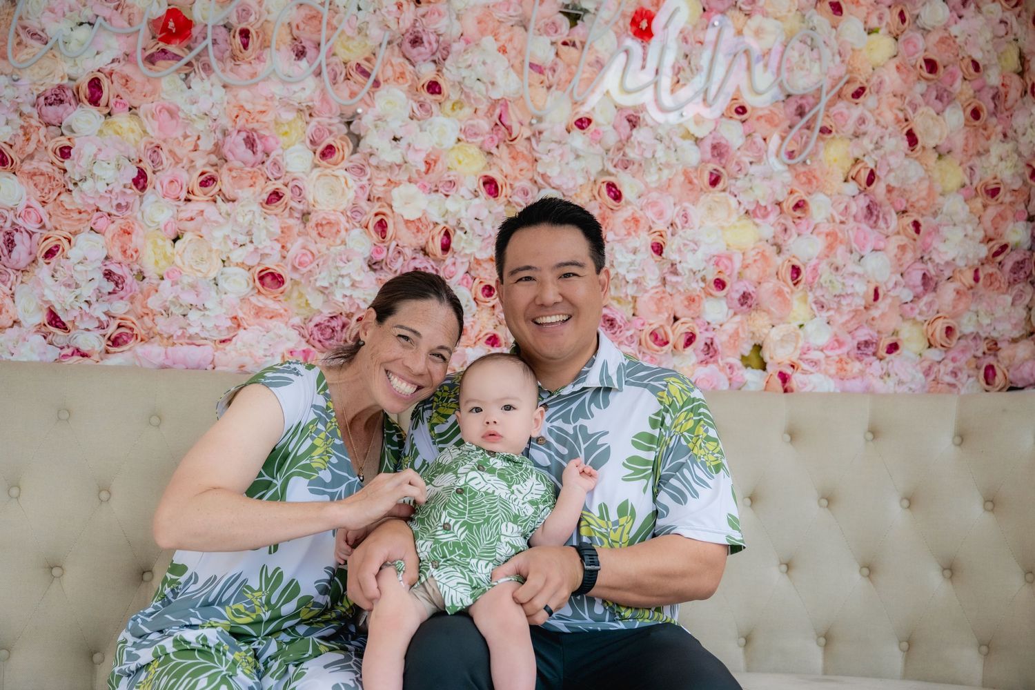 Family of 3 in green aloha print, sitting on white couch in front of wall of paper pink roses.