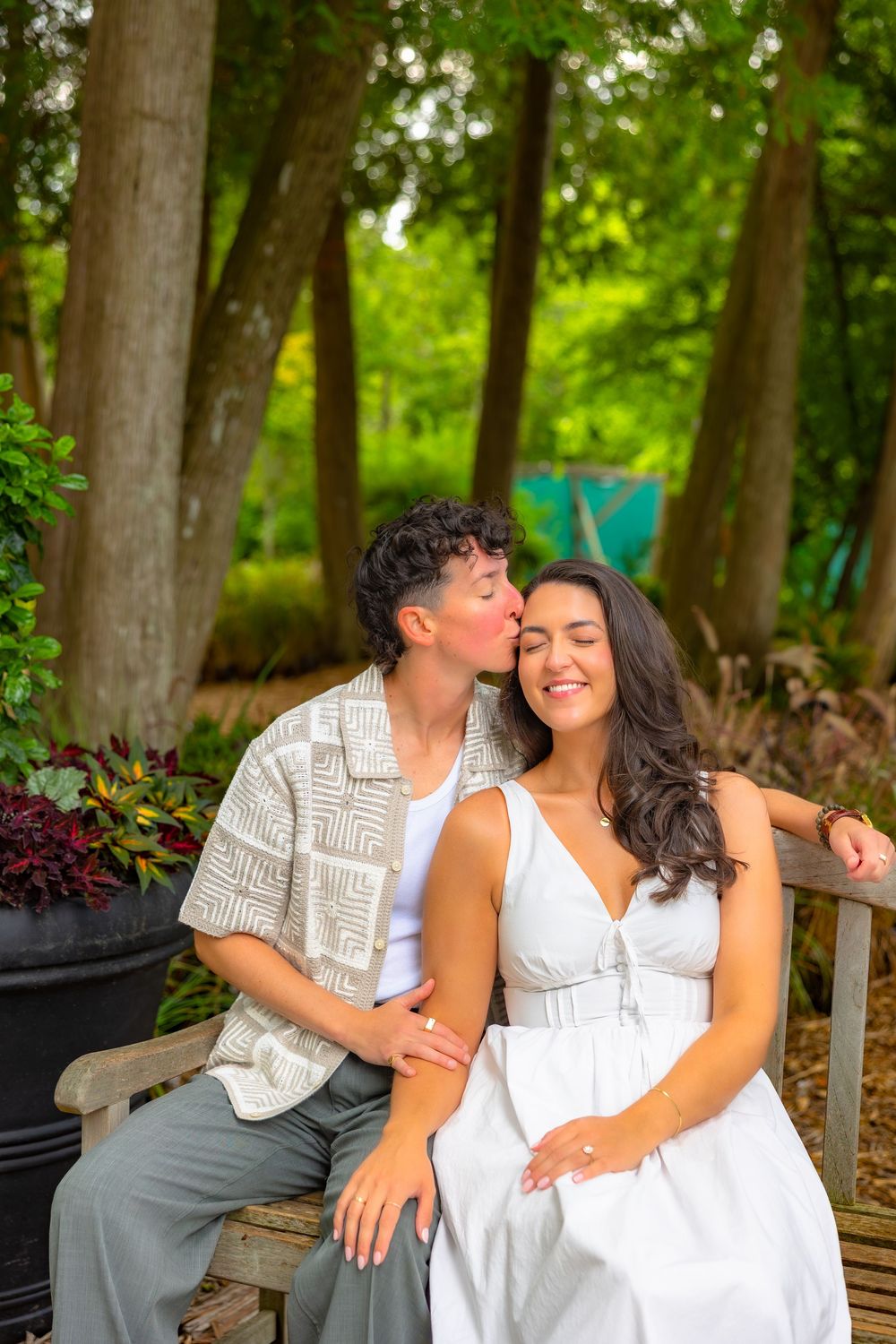 A couple in white outfits share tender moments while sitting on a wooden bench in a lush garden setting.