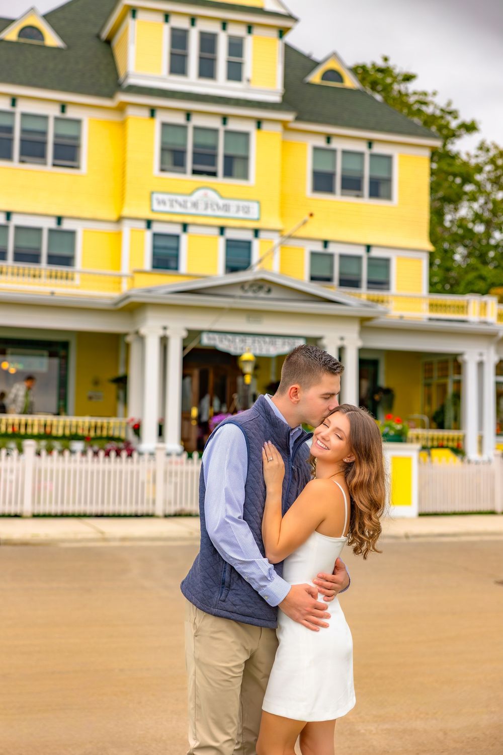 A couple shares a romantic moment in front of a bright yellow beach house with white trim on a sunny day.