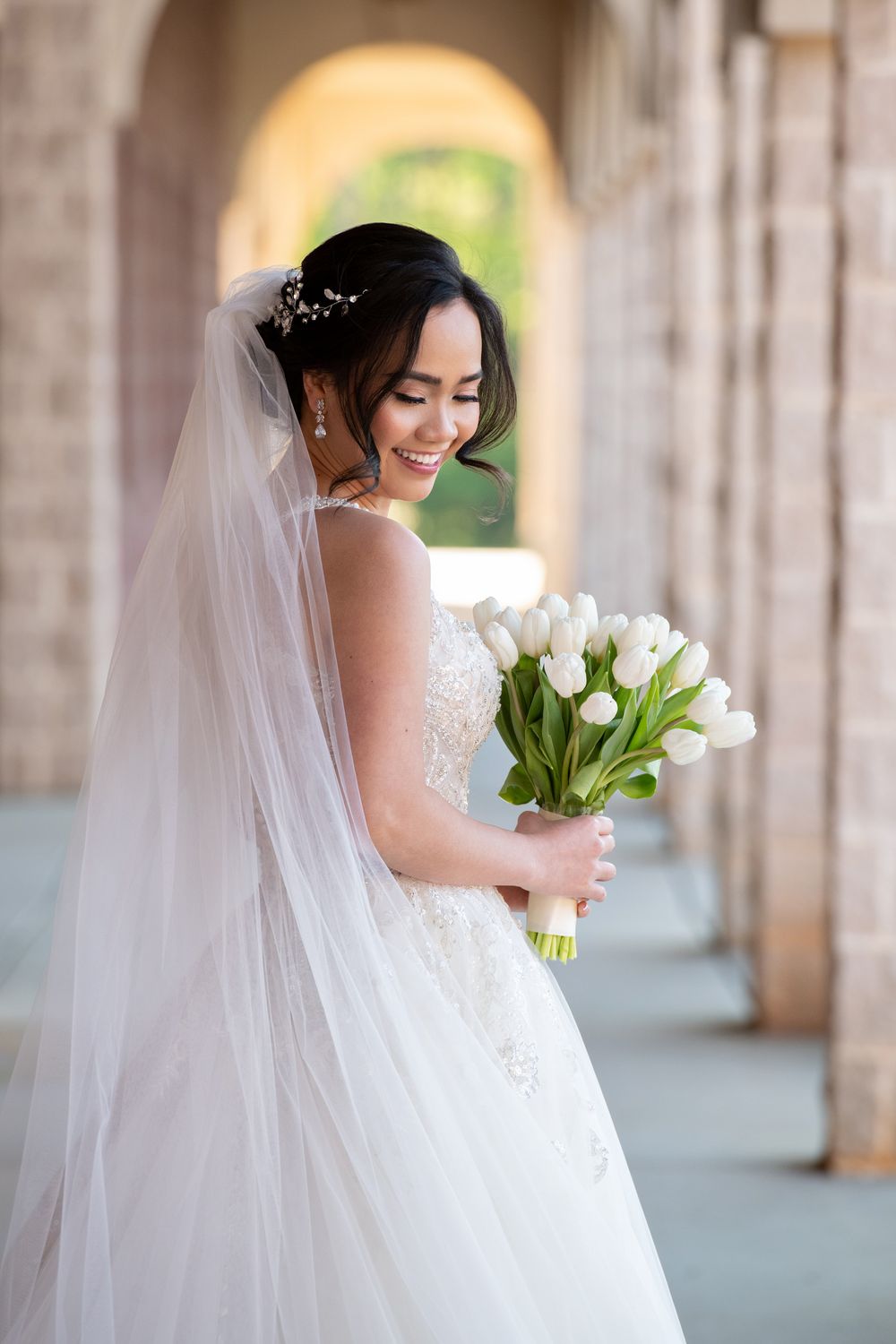 Smiling bride in white lace gown and veil holds white tulip bouquet under arched colonnade.