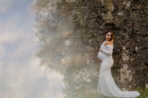 Pregnant woman in white lace gown poses gracefully against a mossy stone wall surrounded by misty trees.
Summerville/Charleston SC