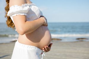 Pregnant woman in white dress standing on beach with ocean in background, cradling bare baby bump.
Folly Beach/Charleston SC