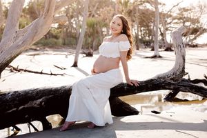 Pregnant woman in white off-shoulder dress sitting on driftwood at beach, smiling during maternity photoshoot.
Folly Beach/Charleston SC