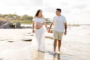 Pregnant couple holding hands walking on beach, woman in white maternity dress, man in white shirt and khaki shorts. Folly Beach/Charleston
