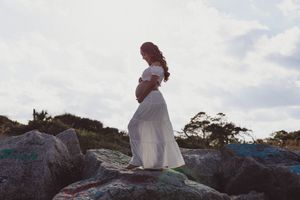 Pregnant woman in white off-shoulder maternity gown standing on rocks with scenic sky backdrop.