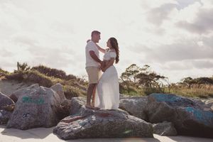 Couple standing on graffiti rocks at a sandy beach, embracing romantically with soft sunlight and coastal vegetation behind them.