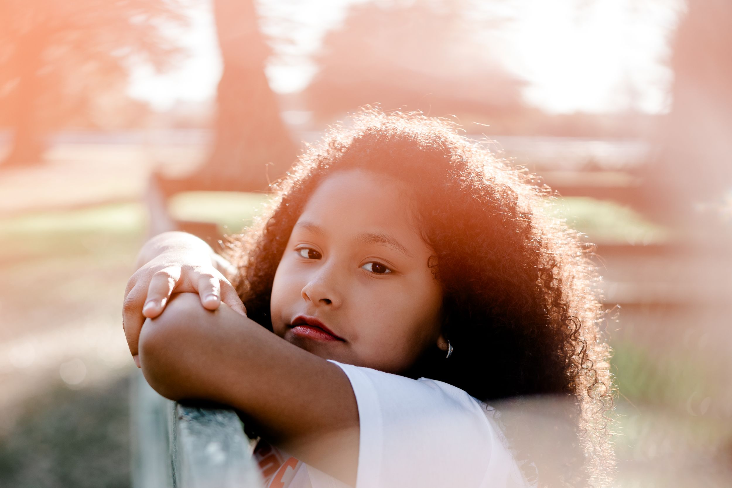 A young girl with curly hair rests her arms on a surface, gazing thoughtfully in soft warm sunlight outdoors.
Summerville/Charleston SC