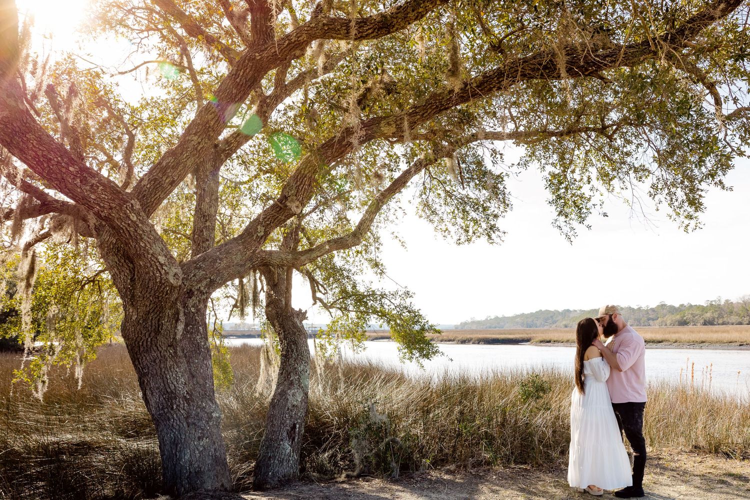 A couple stands under a large oak tree during a romantic sunset photo session in a coastal marsh setting.
