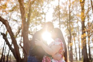 A couple holds hands and walks through a sunlit autumn forest with golden leaves and warm backlighting.