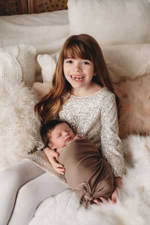 A series of sweet photos showing a young girl in white lace cuddling with a sleeping newborn on soft white bedding.