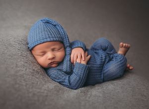 Newborn baby wearing blue knitted outfit sleeping peacefully on gray backdrop in professional studio setting.