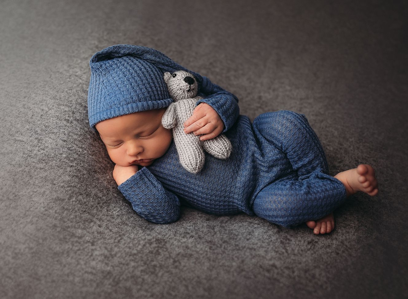 A sleeping newborn in blue knit clothing lies peacefully on a dark gray background in a series of photos.