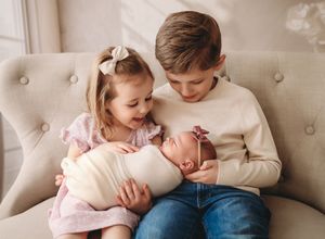 Two young siblings lovingly hold their swaddled newborn baby on a cozy beige sofa.
