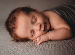 Peaceful newborn baby sleeping on a gray blanket, smiling with rosy cheeks and soft brown hair.