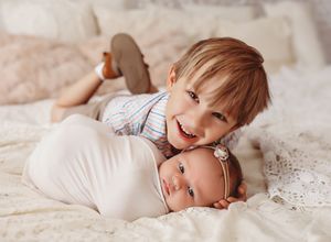 Two siblings share tender moments together while laying on soft white bedding.