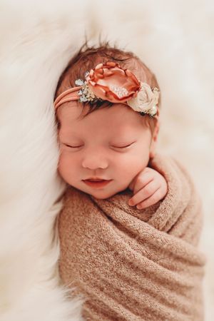 A sleeping newborn wrapped in a beige swaddle blanket wearing a delicate floral headband in a peaceful pose.