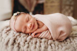 A sleeping newborn baby wrapped in soft pink swaddle blanket lies peacefully on textured white bedding.