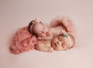 Sleeping newborn baby wearing pink tulle skirt and delicate headband rests peacefully on soft pink background.