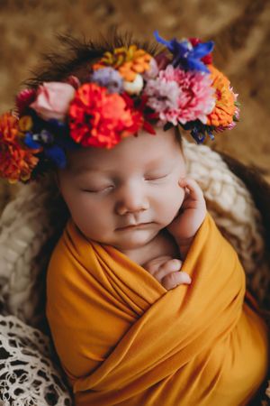 Sleeping baby wrapped in mustard yellow swaddle wearing colorful floral crown with pink, orange and blue blooms.