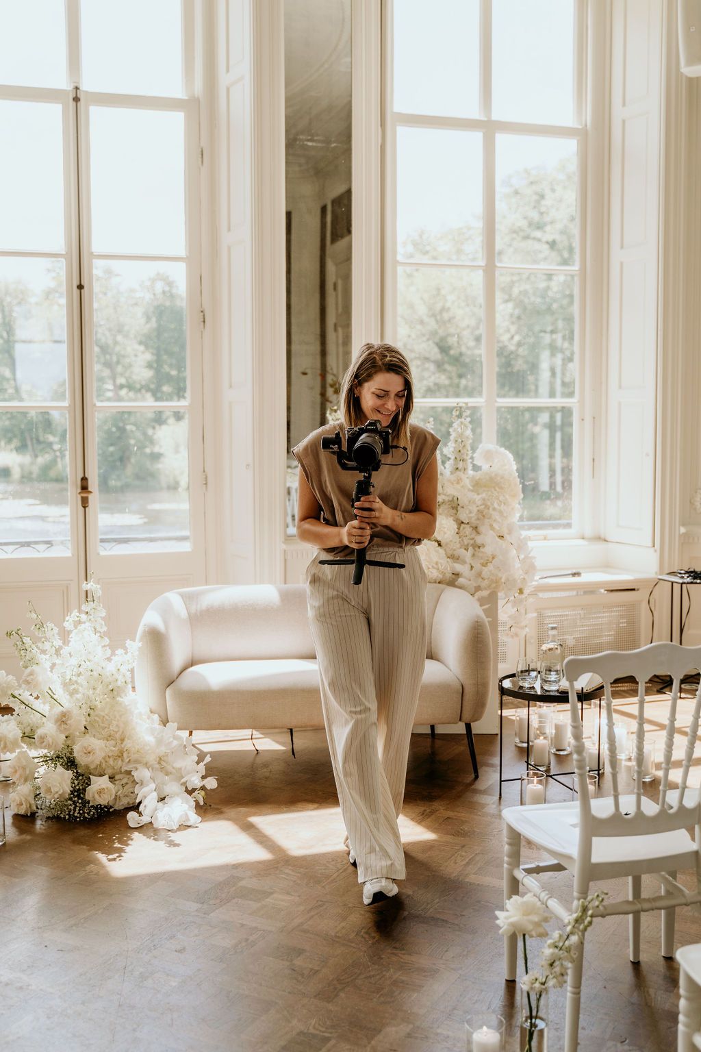 A person in a casual outfit stands in an elegant white room with large windows and floral decorations.