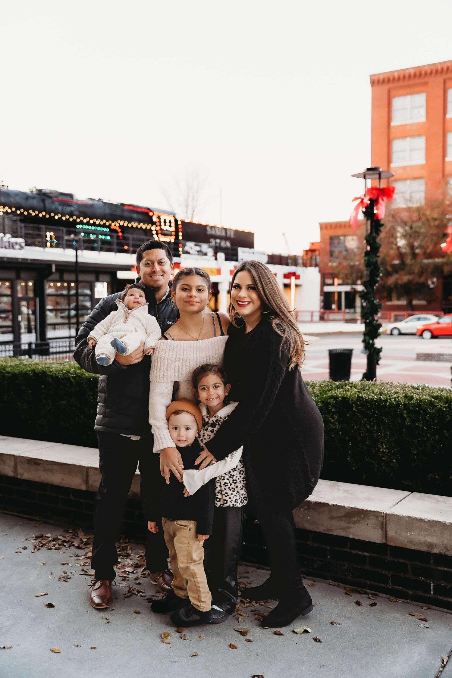 A family poses together on a sidewalk during the holiday season with decorative lights and buildings in the background.