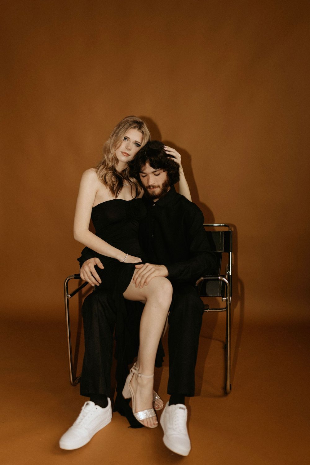 Stylish couple in black clothing pose intimately on a chair against warm brown background.