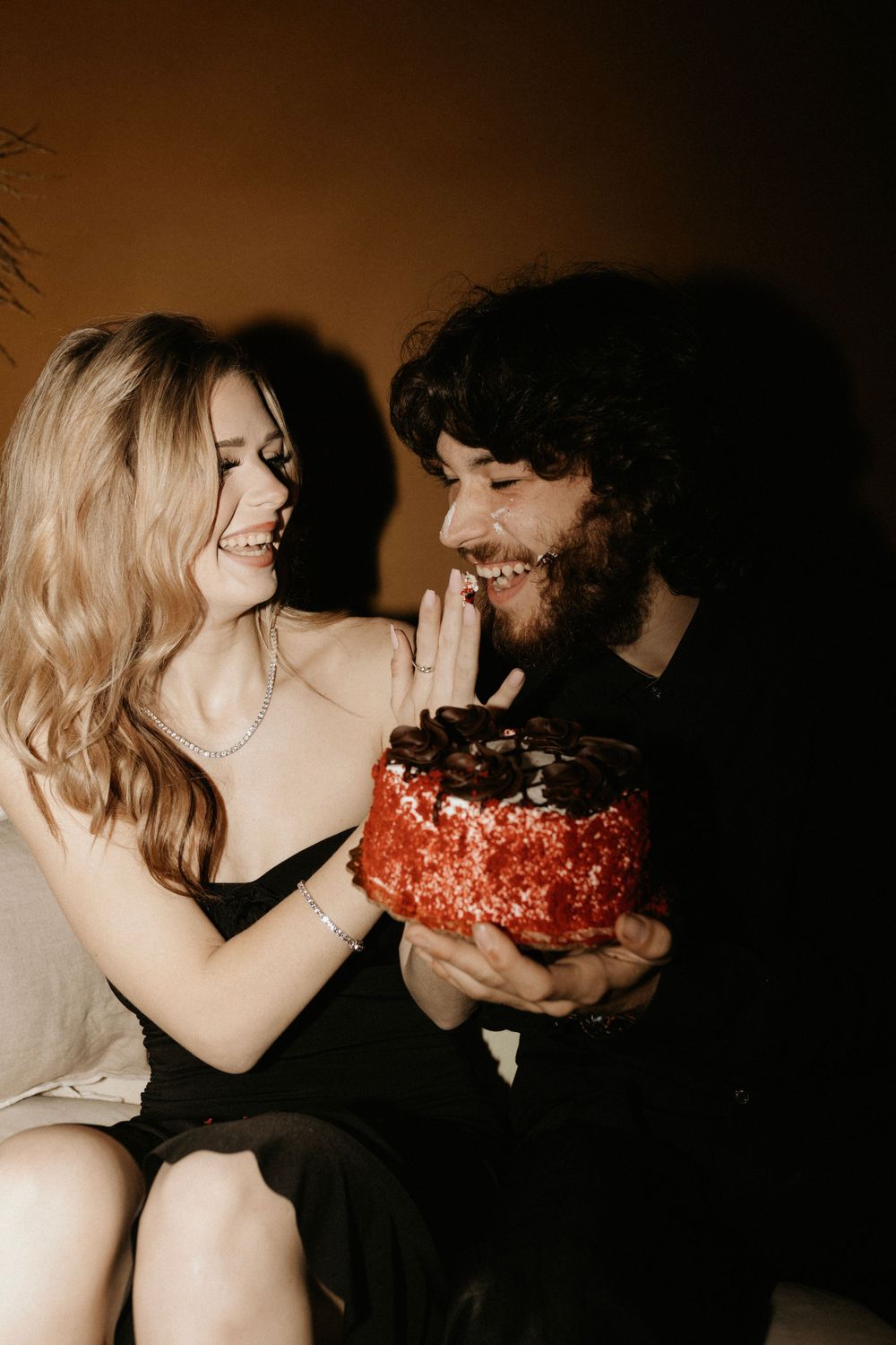 Couple sharing a playful moment with red birthday cake against dark background.