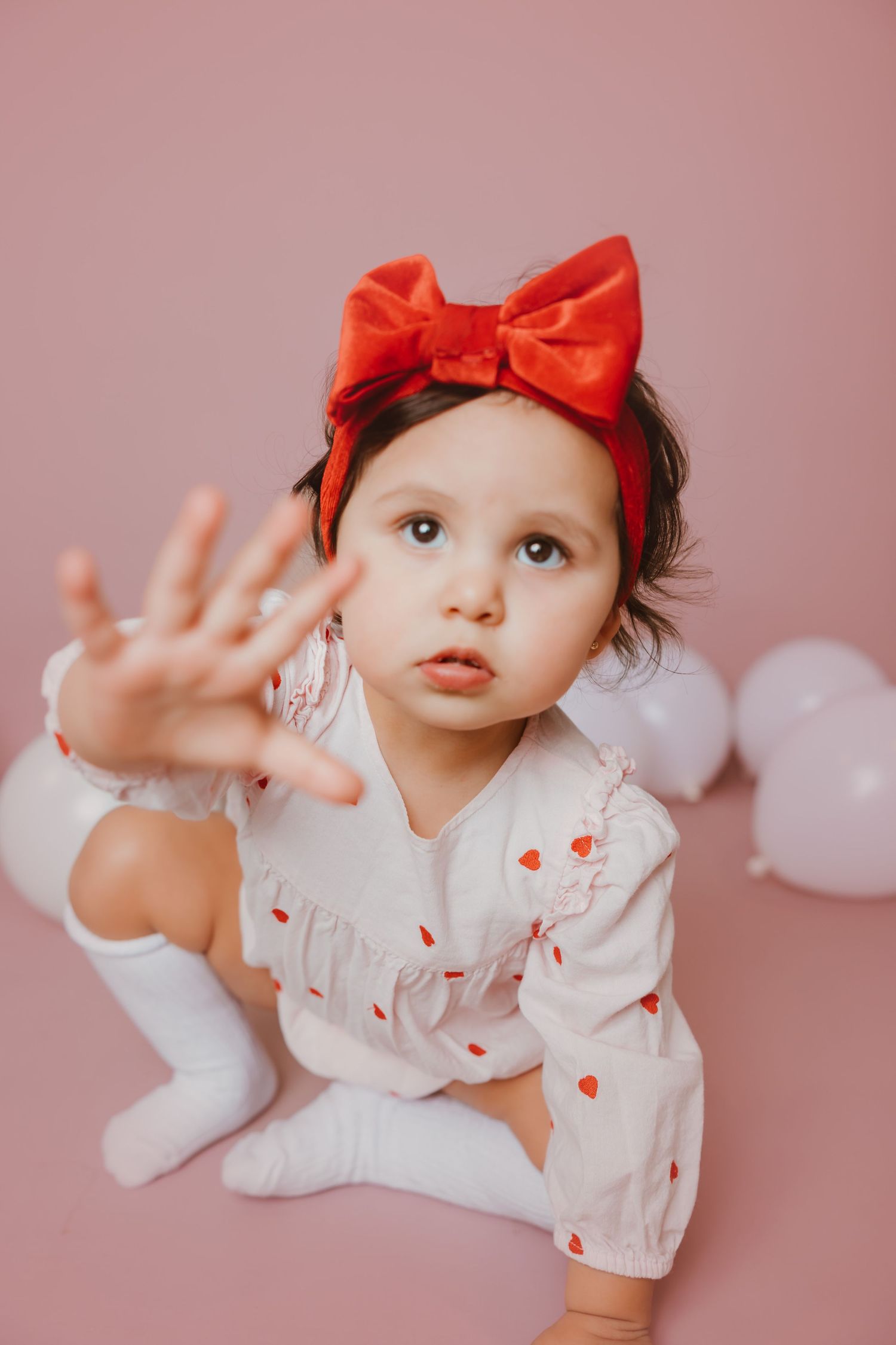 Toddler in white dress with red bow headband reaching toward camera on pink background.