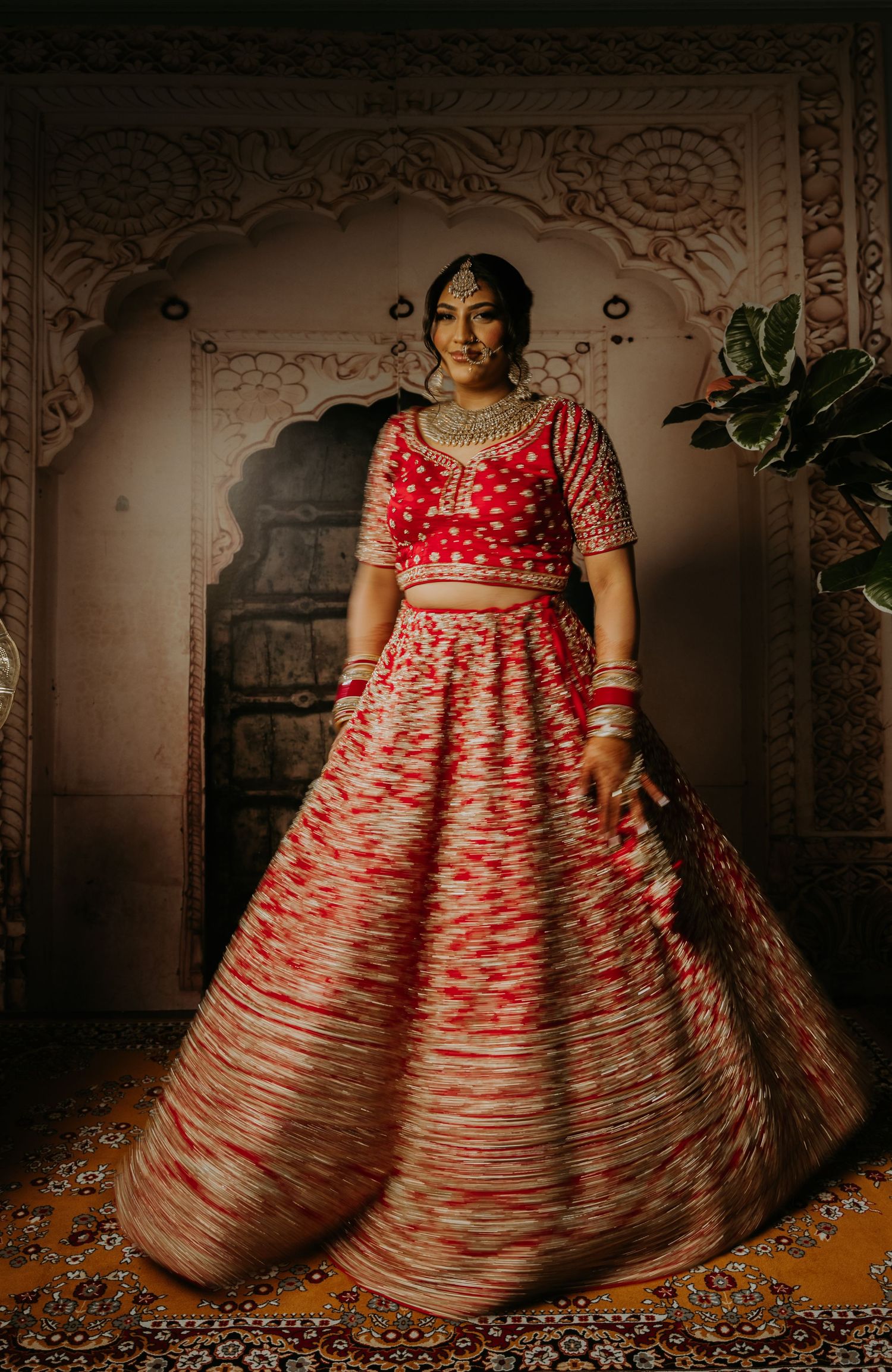 Traditional Indian bride in red and white lehenga poses against ornate architectural backdrop.