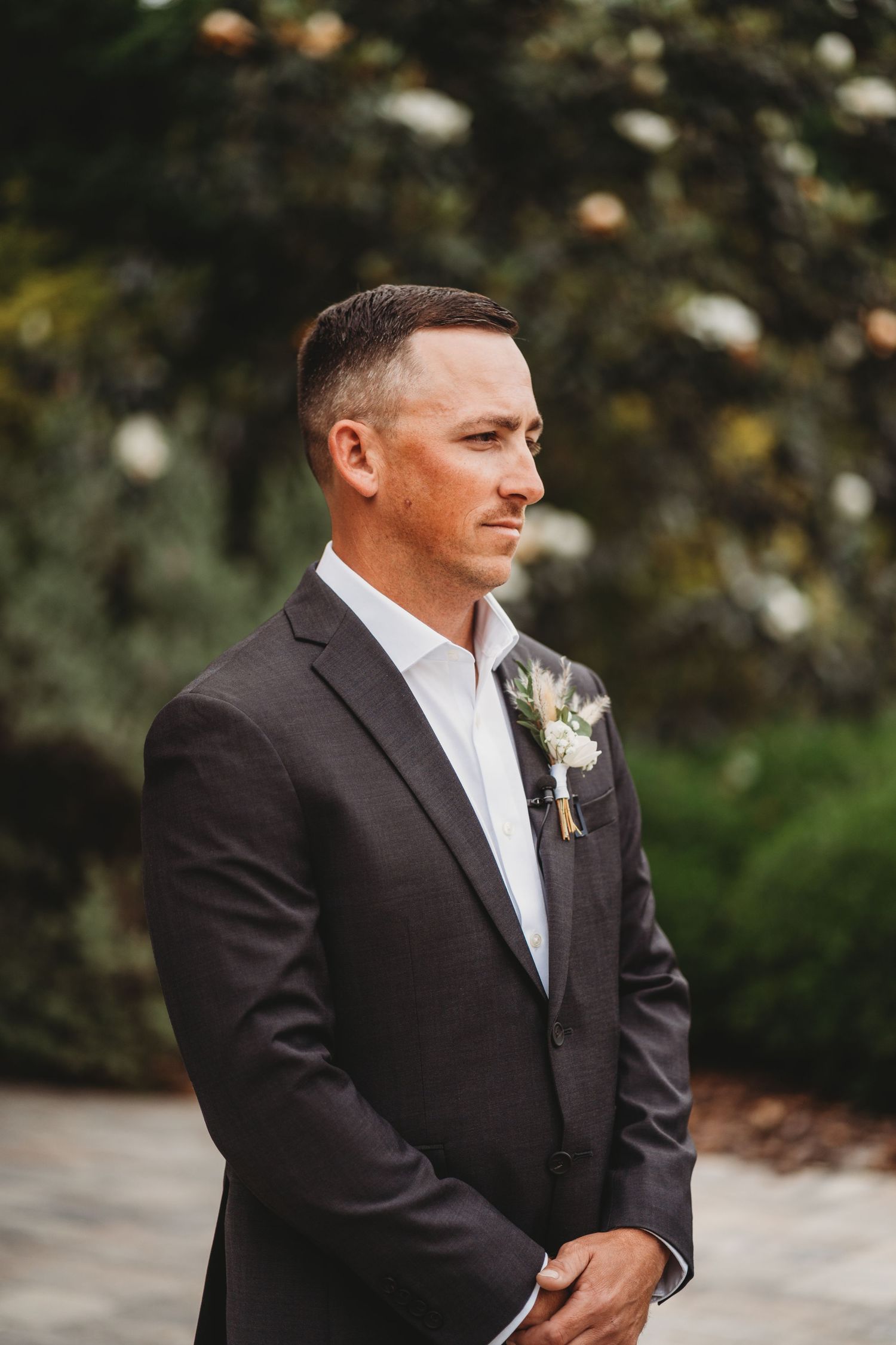 Man in dark suit with boutonniere standing outdoors during wedding ceremony.