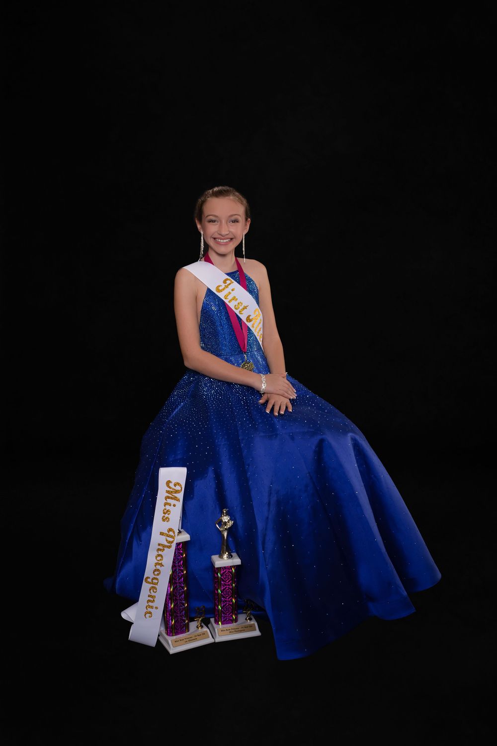 Pageant contestant in royal blue ball gown poses with sash and trophy against black background.