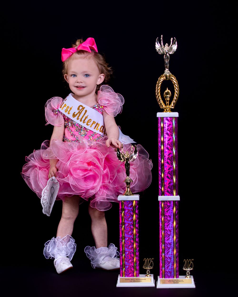 A toddler in a sparkling pink pageant dress and glittering crown poses next to dance competition trophies.
