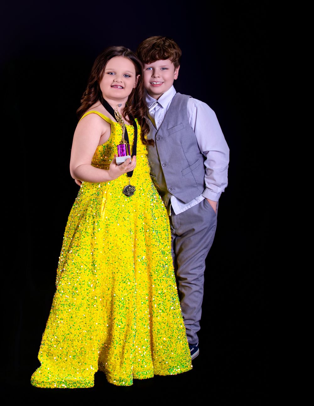 Two people pose together at a formal event, with a bright yellow sequined ballgown and grey suit against a dark backdrop.