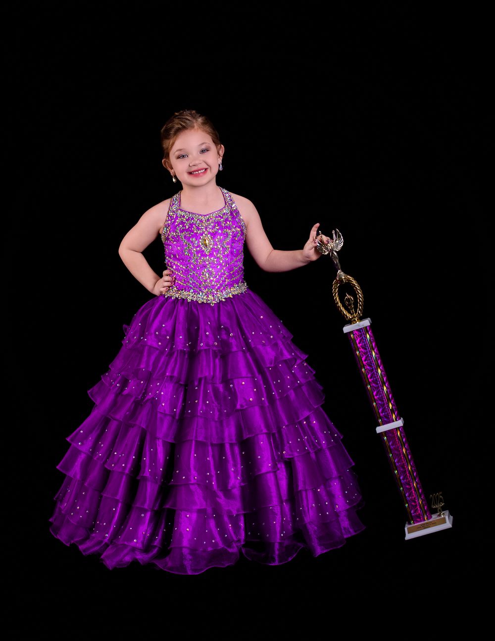 A child in a sparkly purple pageant gown with tiered ruffles poses with a trophy on a black background.