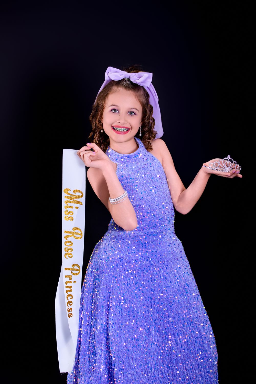 Young contestant in a sparkly purple gown and hair bow poses with pageant sash and crown against dark background.