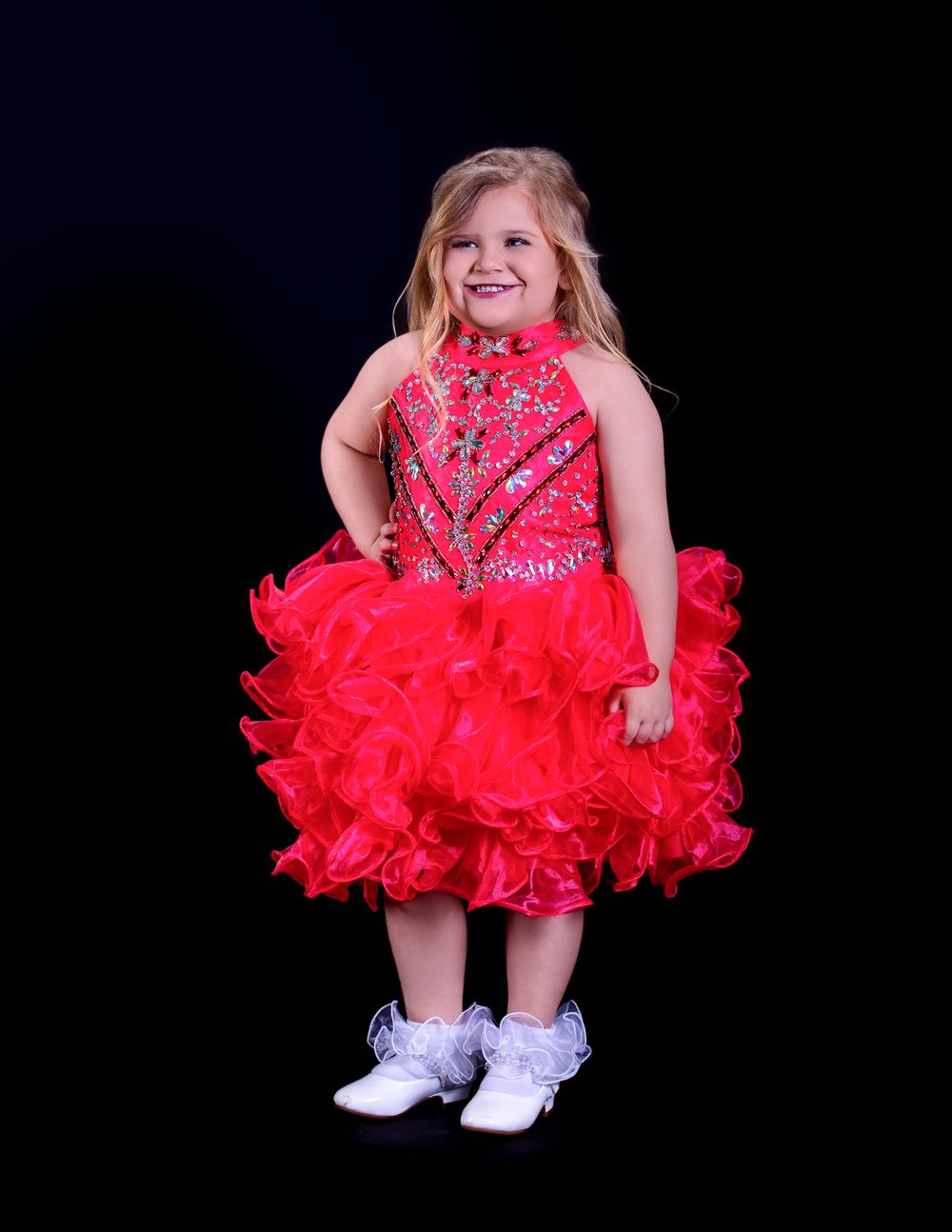 A young dancer in a red ruffled pageant dress with rhinestones poses against a black background for portraits.