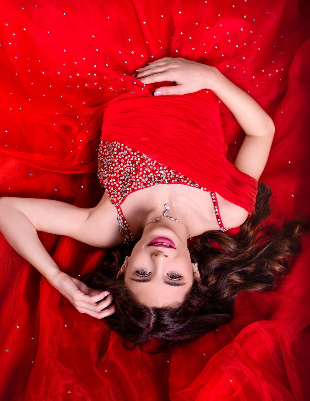 Woman in red evening gown with rhinestone details lying on red fabric backdrop looking upward with glamorous makeup.