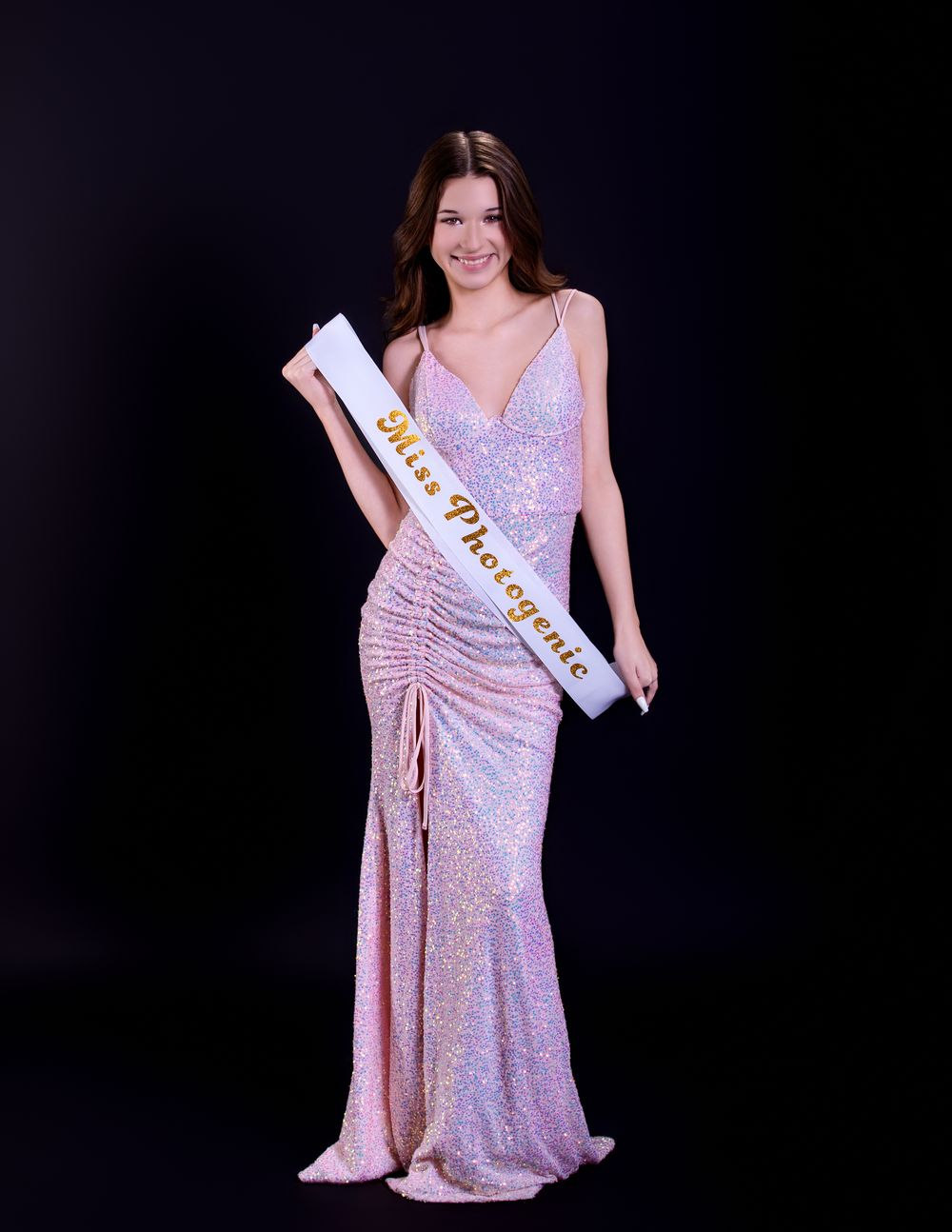 A beauty pageant contestant wearing a sparkly pink evening gown and sash poses against a black backdrop.