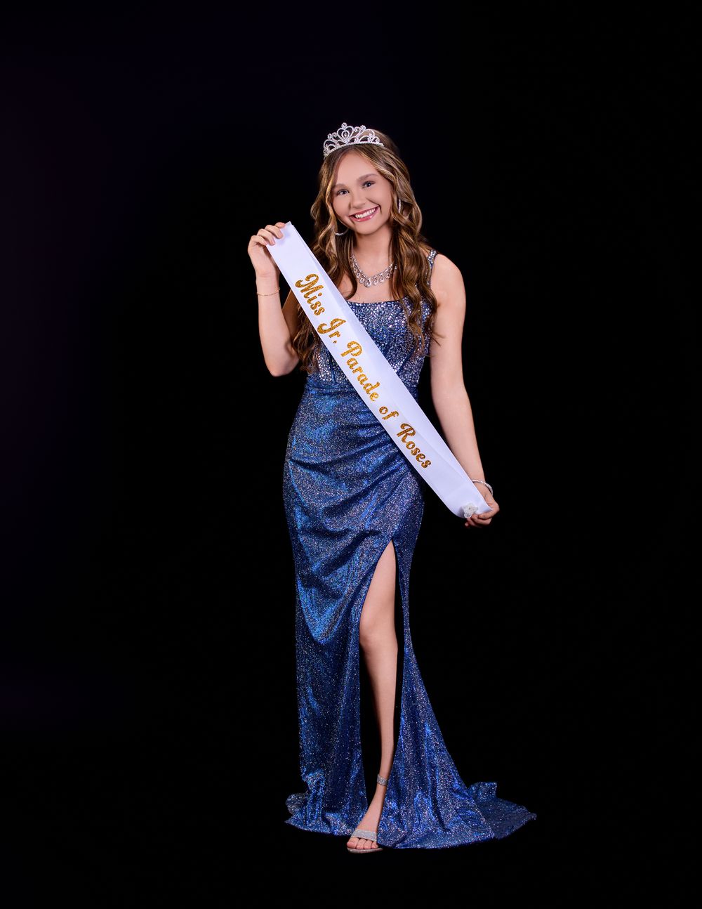 A pageant contestant in a sparkly blue evening gown and tiara poses with a sash against a black background.