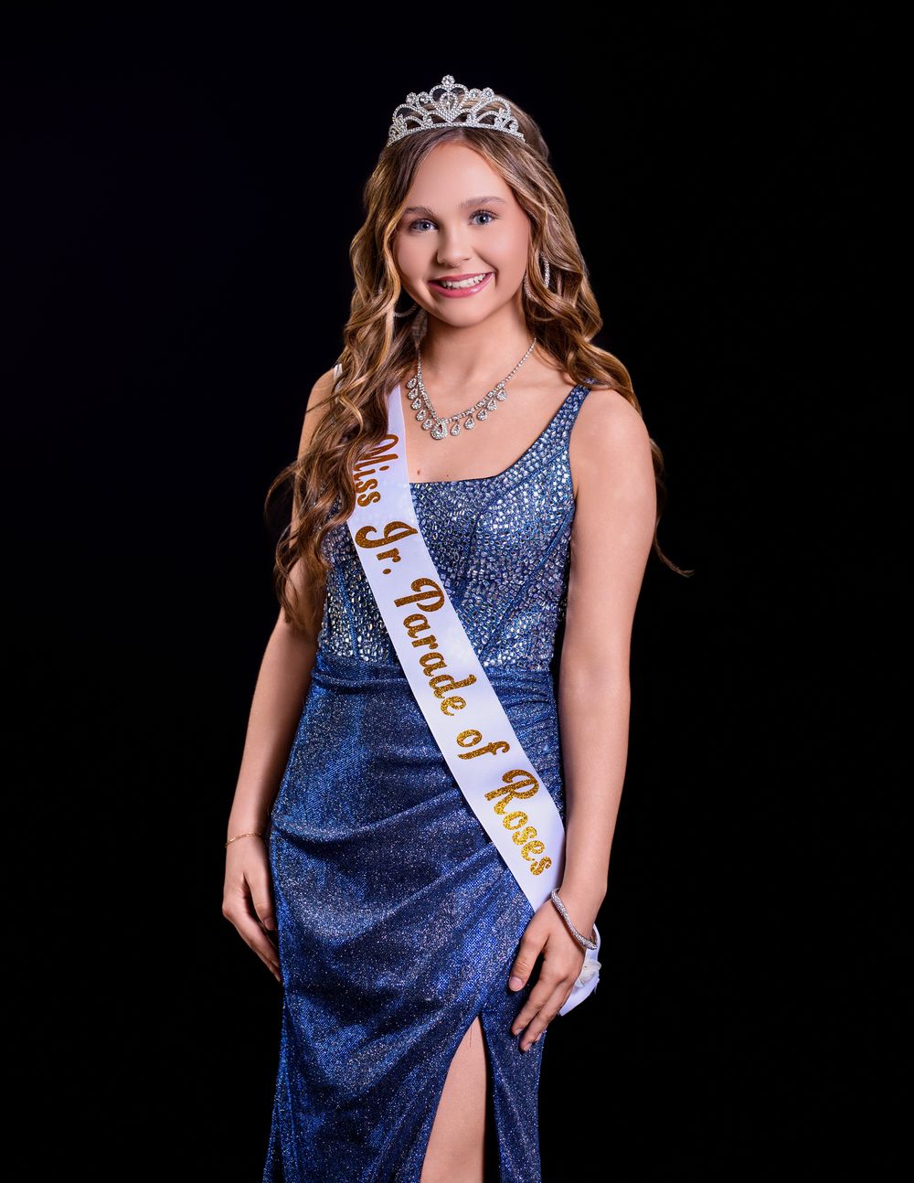 Figure in a sparkling navy blue evening gown and tiara poses for a formal portrait against a black backdrop.