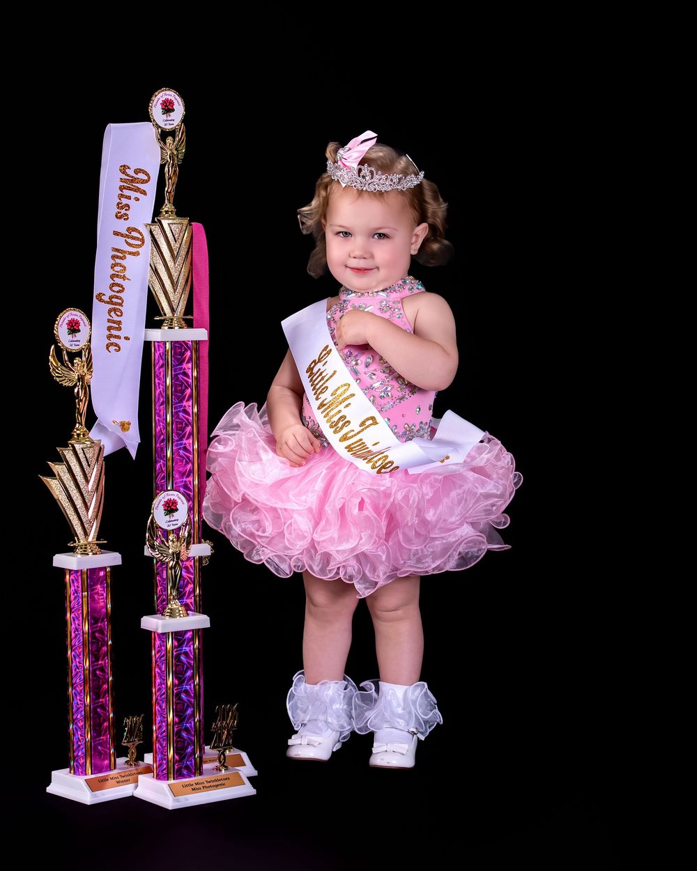 Toddler girl in pink tutu dress and tiara poses with Miss Photogenic pageant trophies.