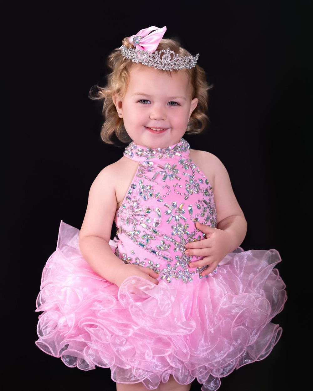 Toddler girl in pink rhinestone pageant dress and tiara smiling against black background.