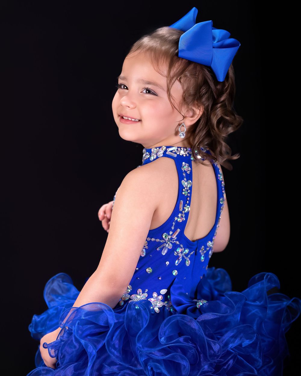 Young girl in royal blue pageant dress with bow hair accessory, smiling over shoulder against dark background.