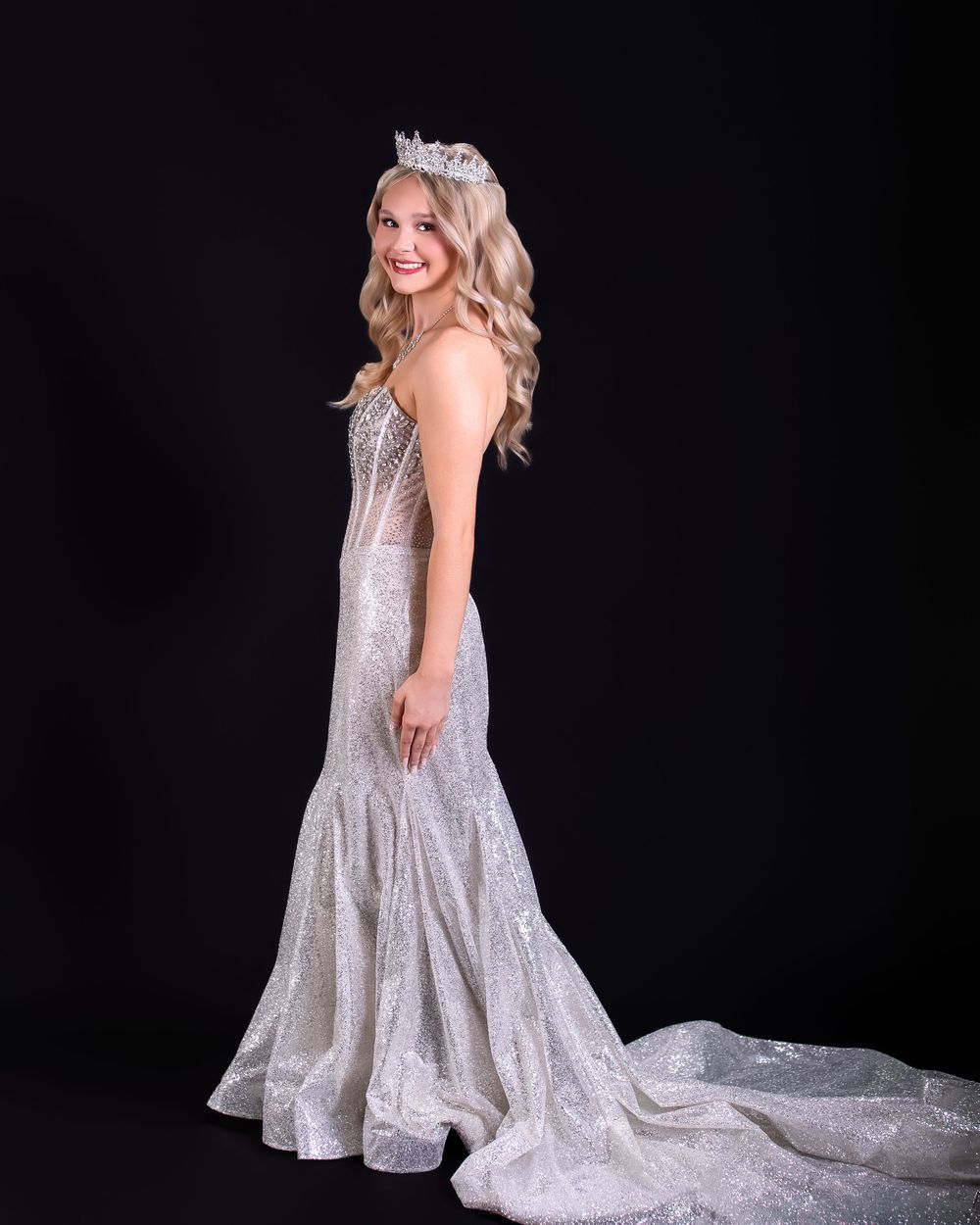 Young woman in sparkling silver mermaid gown and rhinestone crown poses gracefully against black background.