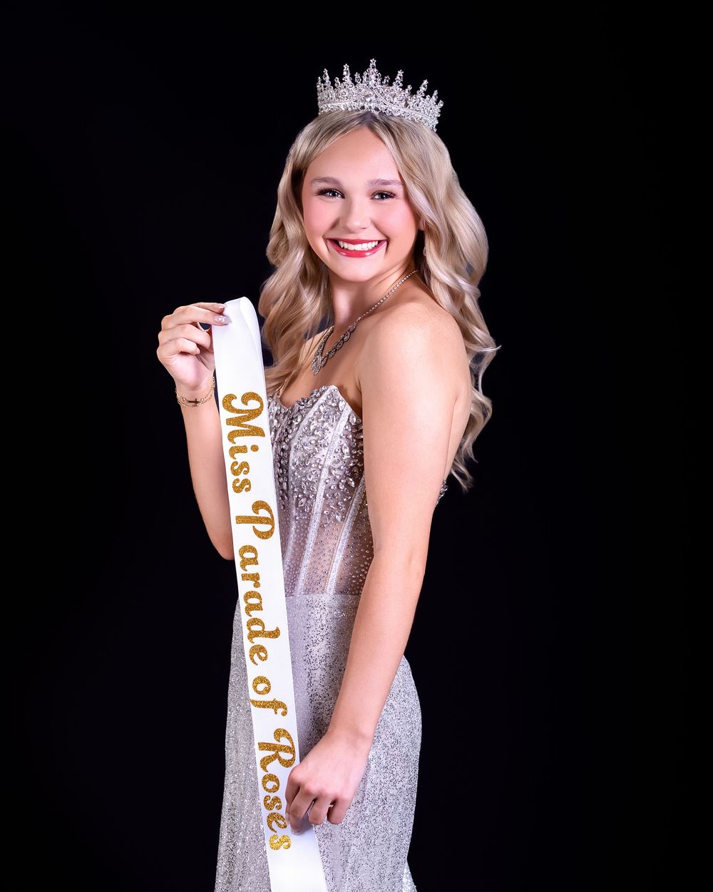 Young woman in sparkly gown wearing Miss Parade of Roses sash and crown poses against black background.